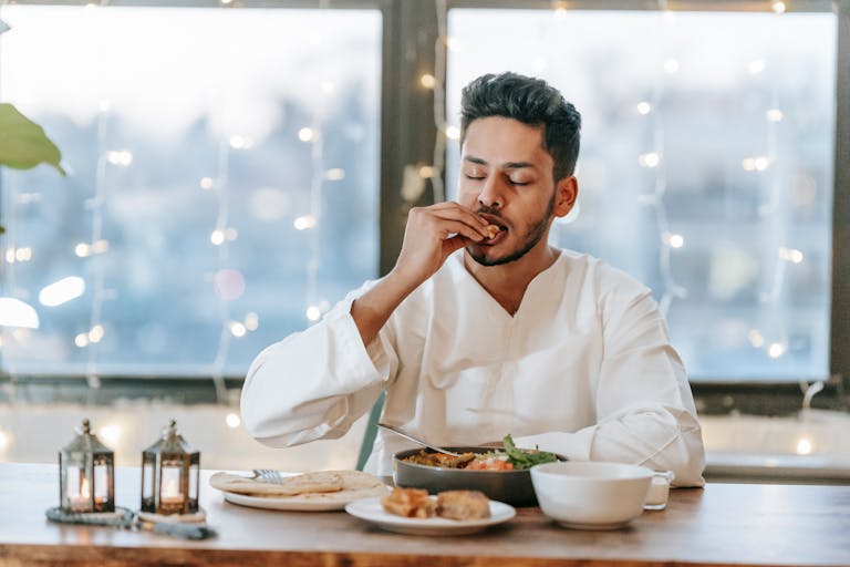 A man in a white thobe savoring a traditional meal indoors with string lights background.