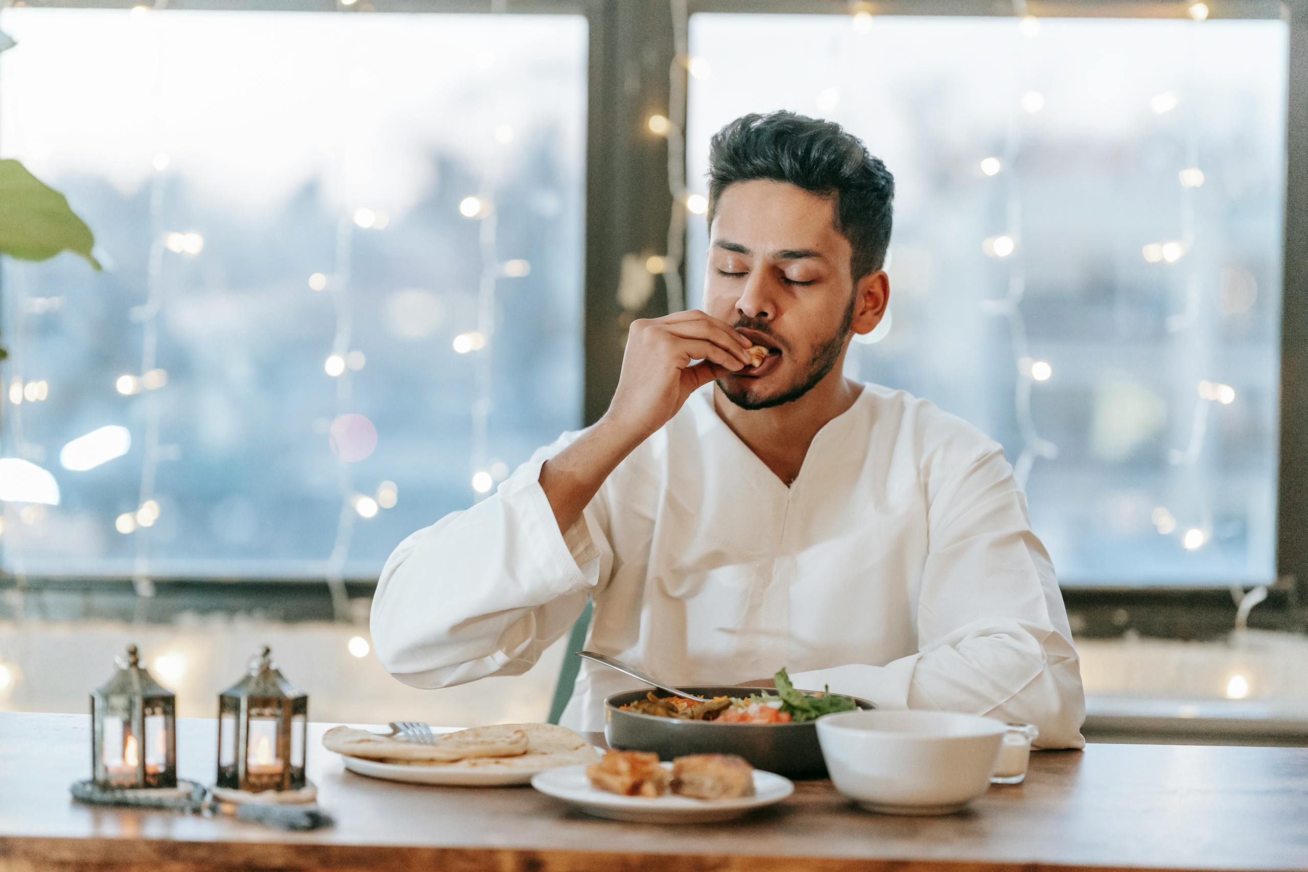 A man in a white thobe savoring a traditional meal indoors with string lights background.