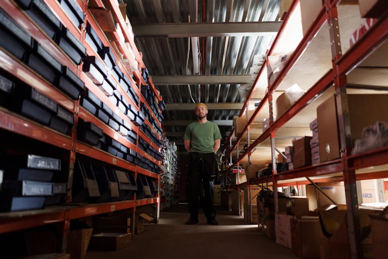 A man standing in a warehouse aisle surrounded by shelves filled with boxes and inventory.