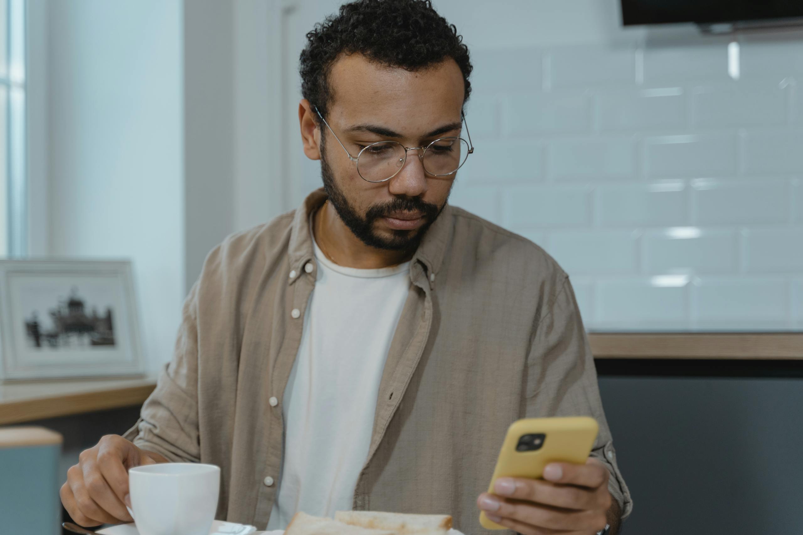 A man with eyeglasses using a smartphone while enjoying breakfast indoors. Perfect for lifestyle and technology themes.