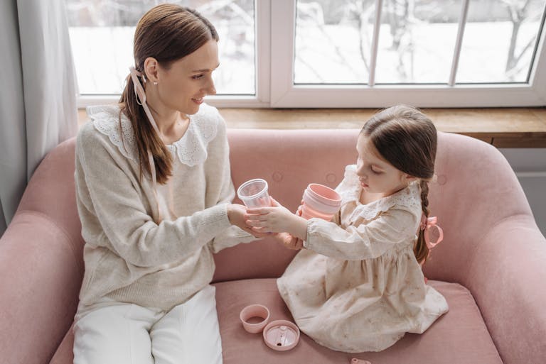 A mother and daughter share a playful moment on a pink sofa with plastic cups indoors.