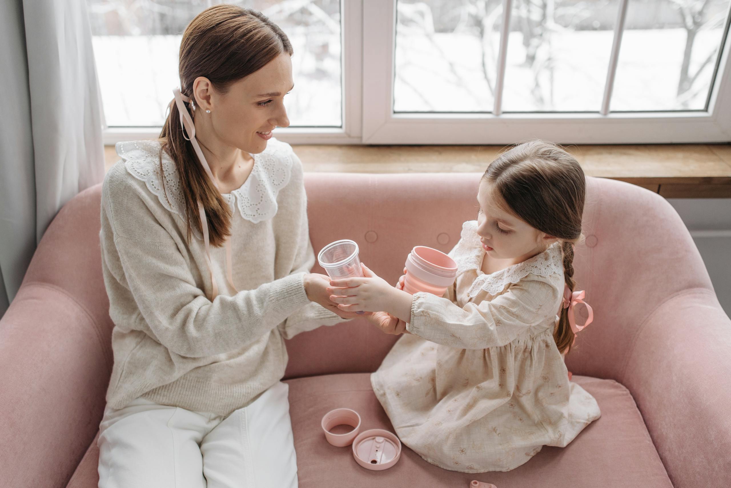 A mother and daughter share a playful moment on a pink sofa with plastic cups indoors.
