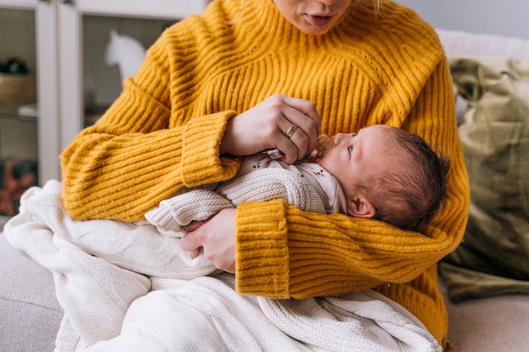 A mother lovingly holds her newborn baby, wrapped in a blanket, wearing a yellow sweater indoors.