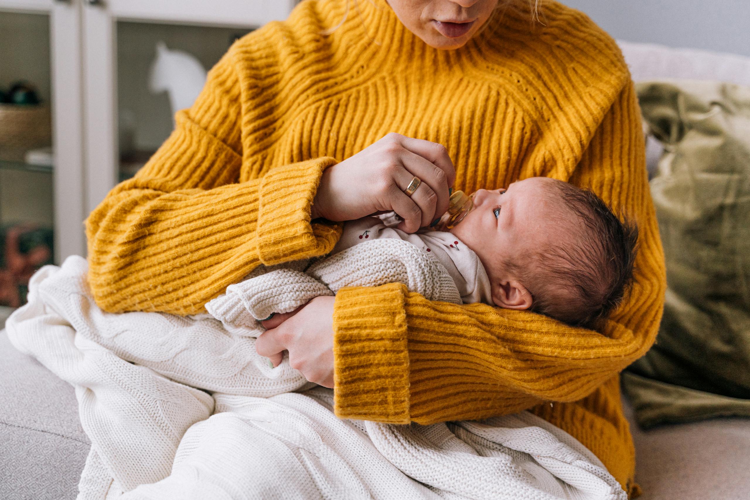 A mother lovingly holds her newborn baby, wrapped in a blanket, wearing a yellow sweater indoors.
