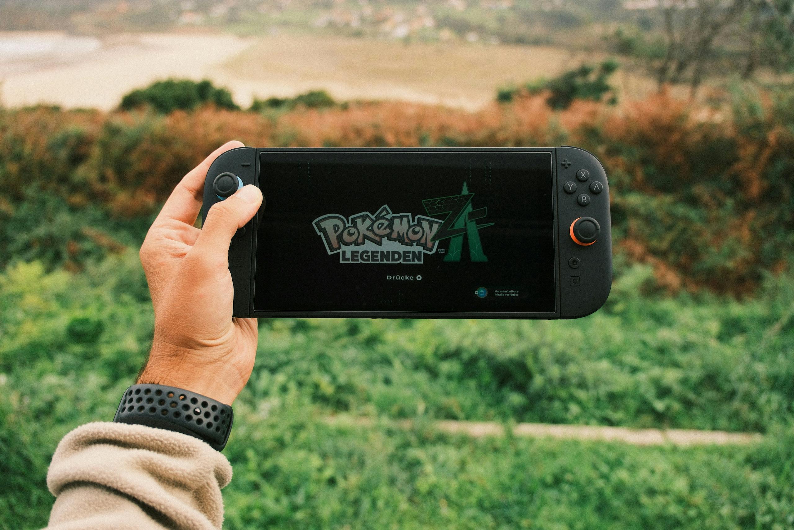 A person holds a handheld gaming device outdoors with Pokémon Legends on screen.