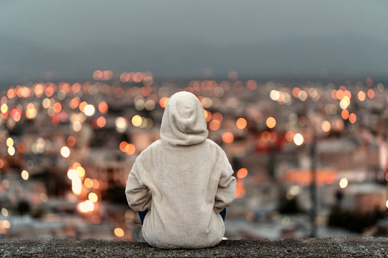A person in a white hoodie sits on a ledge overlooking a cityscape with bokeh lights.