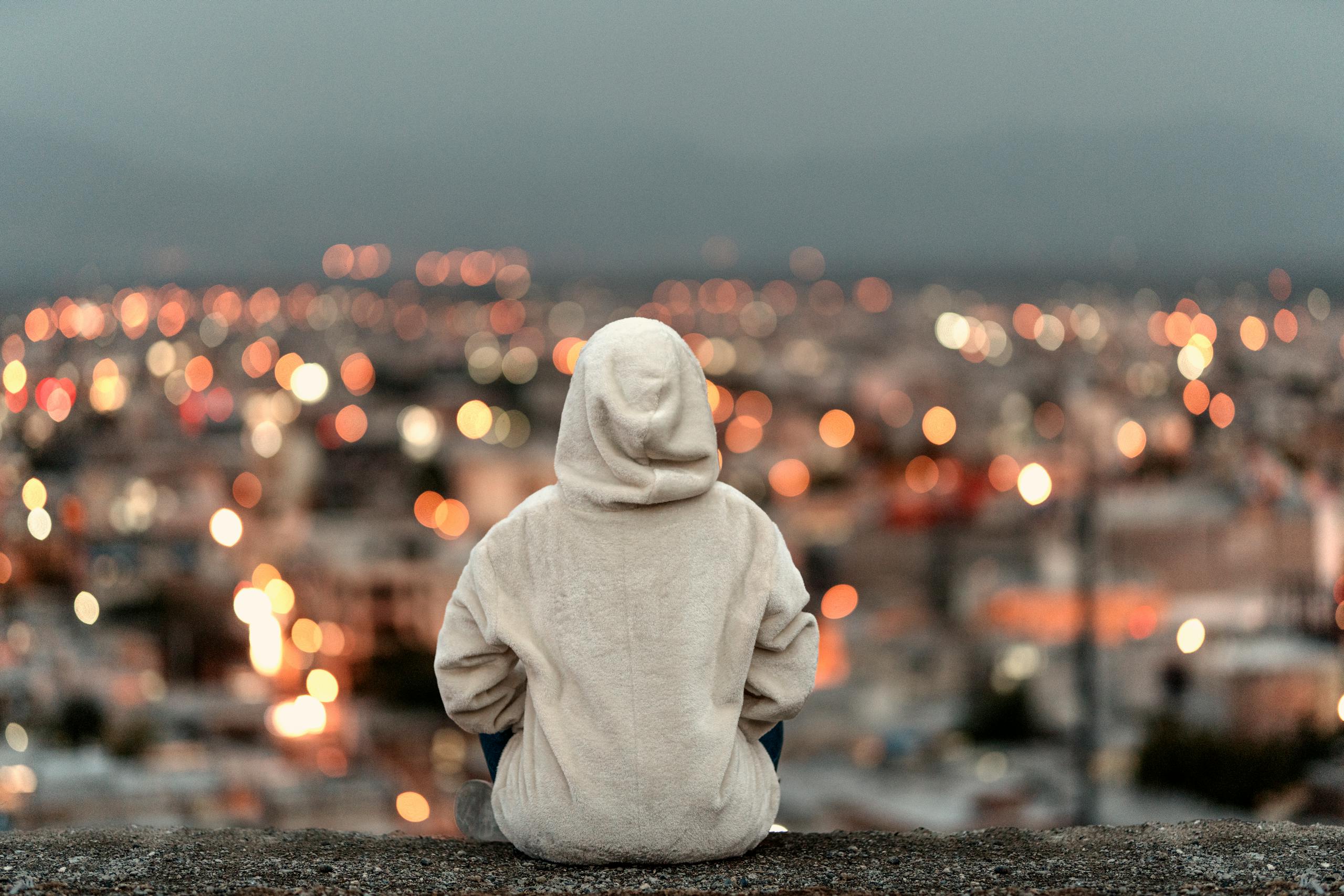 A person in a white hoodie sits on a ledge overlooking a cityscape with bokeh lights.