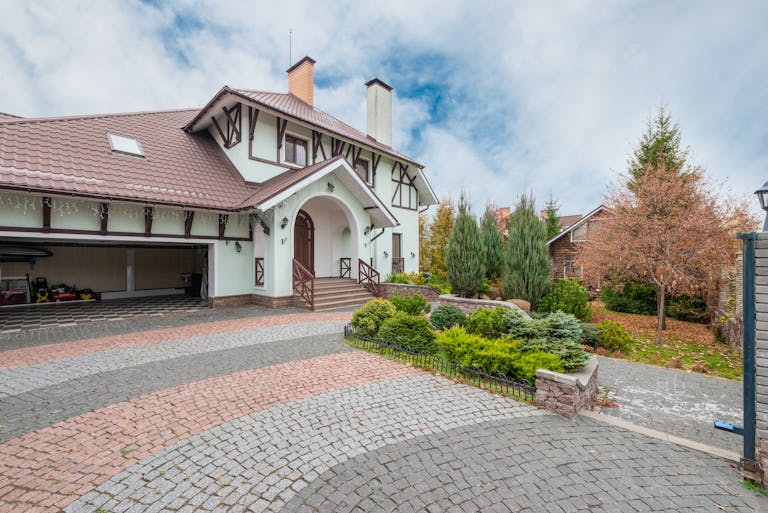 A quaint suburban house with a beautiful garden and brick driveway under a partly cloudy sky.
