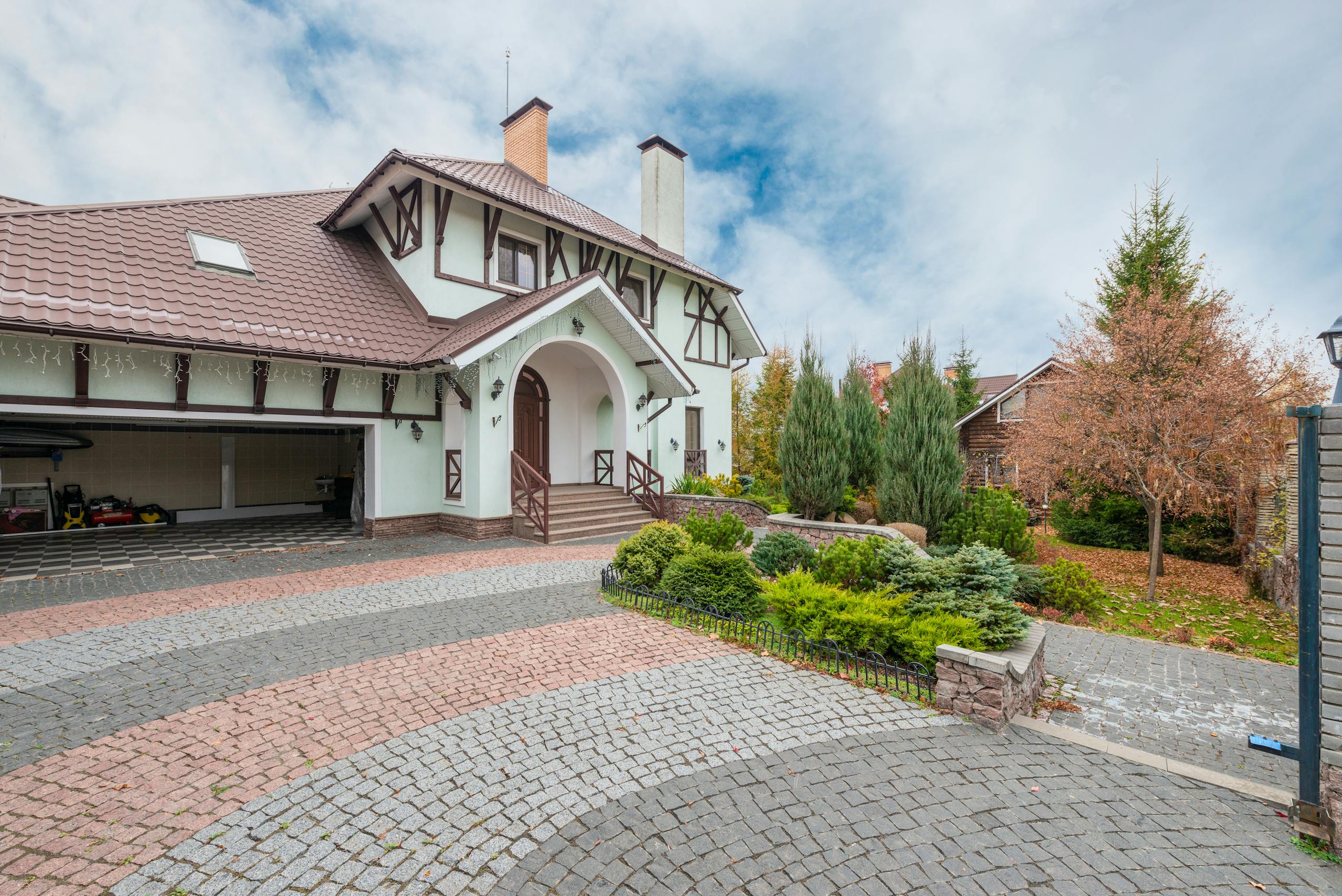 A quaint suburban house with a beautiful garden and brick driveway under a partly cloudy sky.