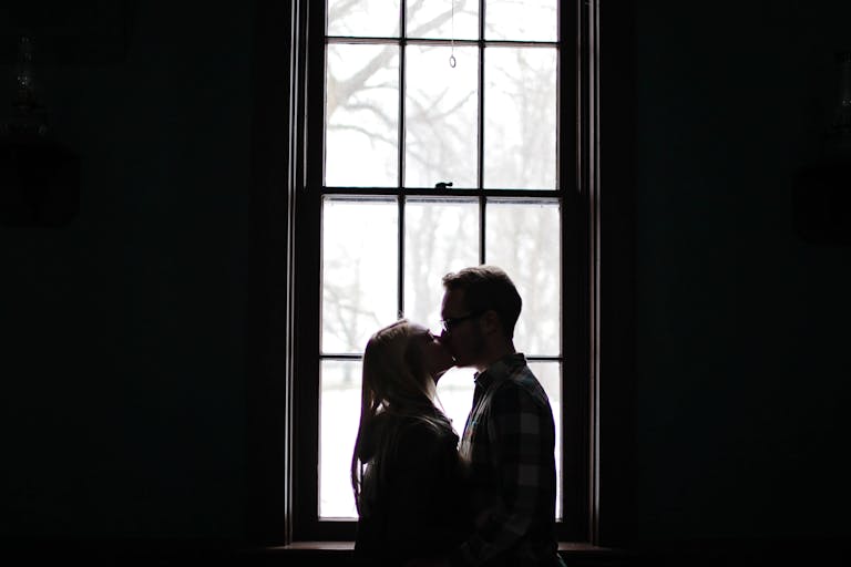 A romantic silhouette of a couple sharing a kiss in front of a large, backlit window.