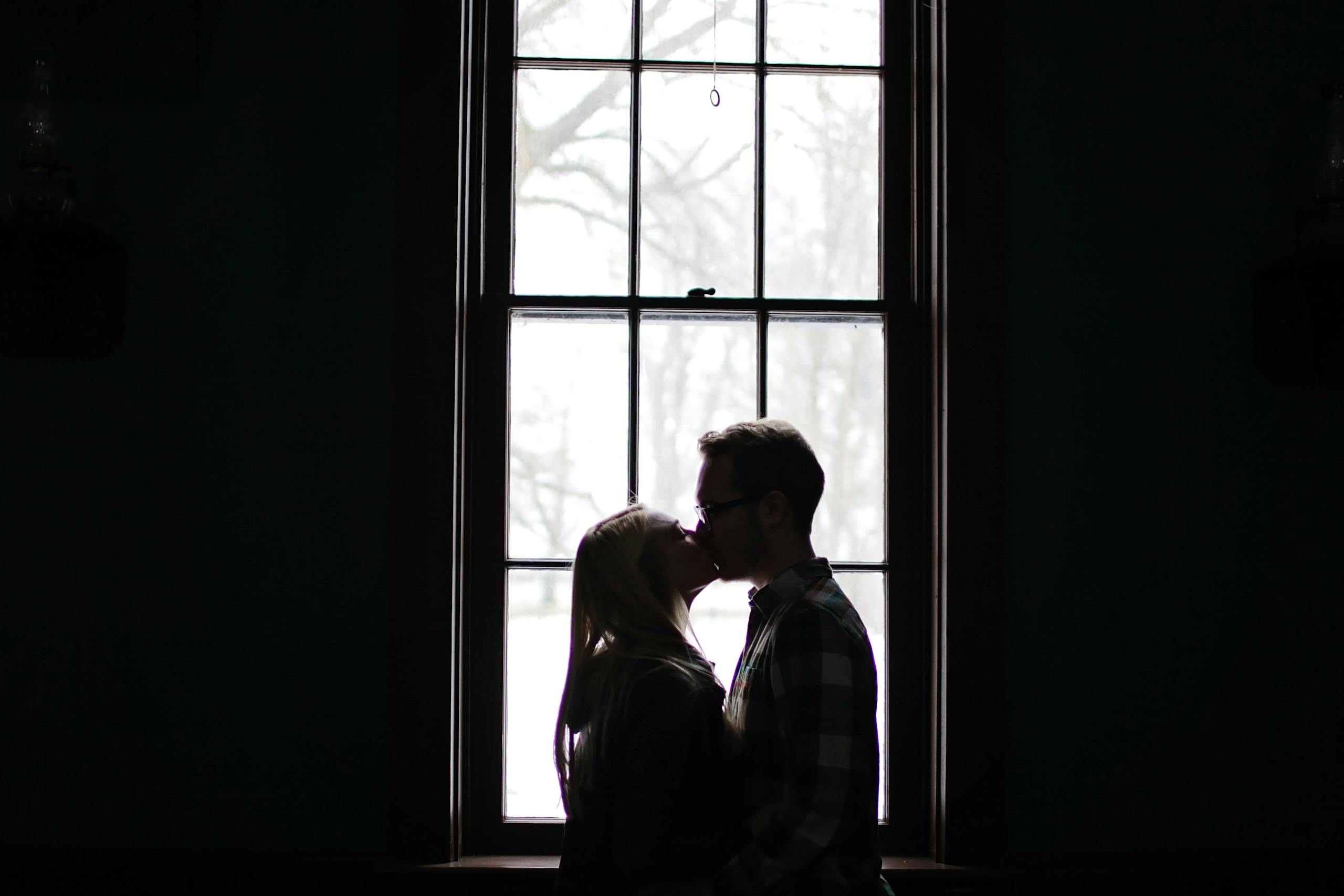 A romantic silhouette of a couple sharing a kiss in front of a large, backlit window.