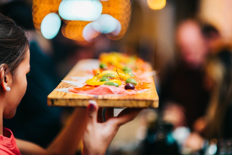 A server presents a vibrant appetizer platter with olives and peppers in a lively restaurant setting.
