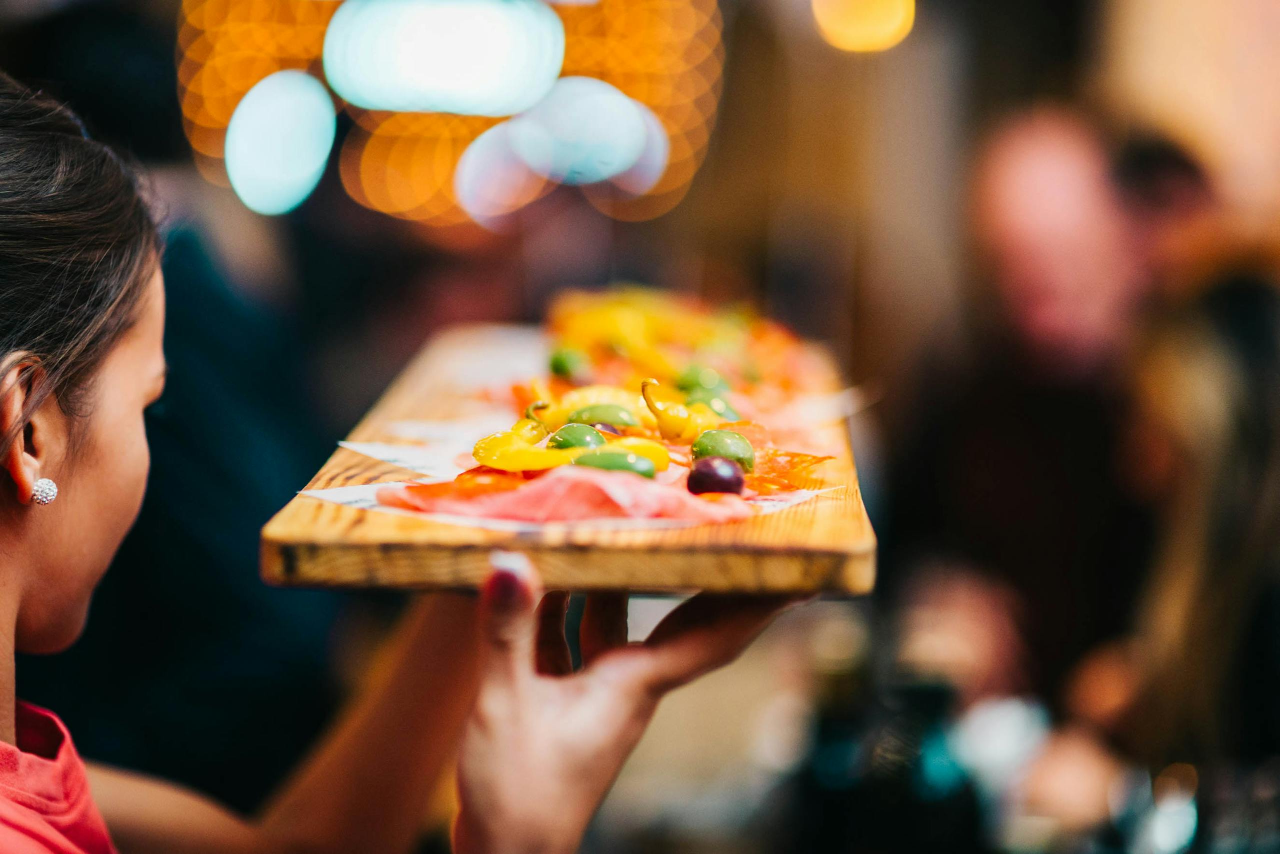A server presents a vibrant appetizer platter with olives and peppers in a lively restaurant setting.