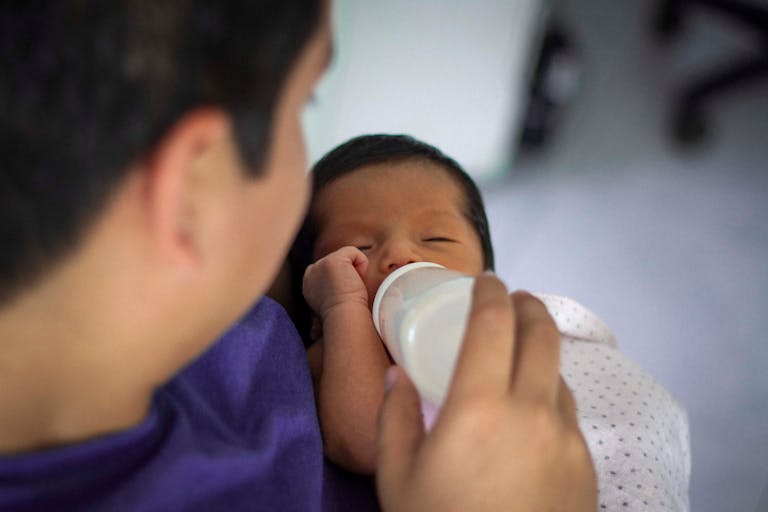 A tender moment captured as a father feeds his newborn baby with a bottle indoors.