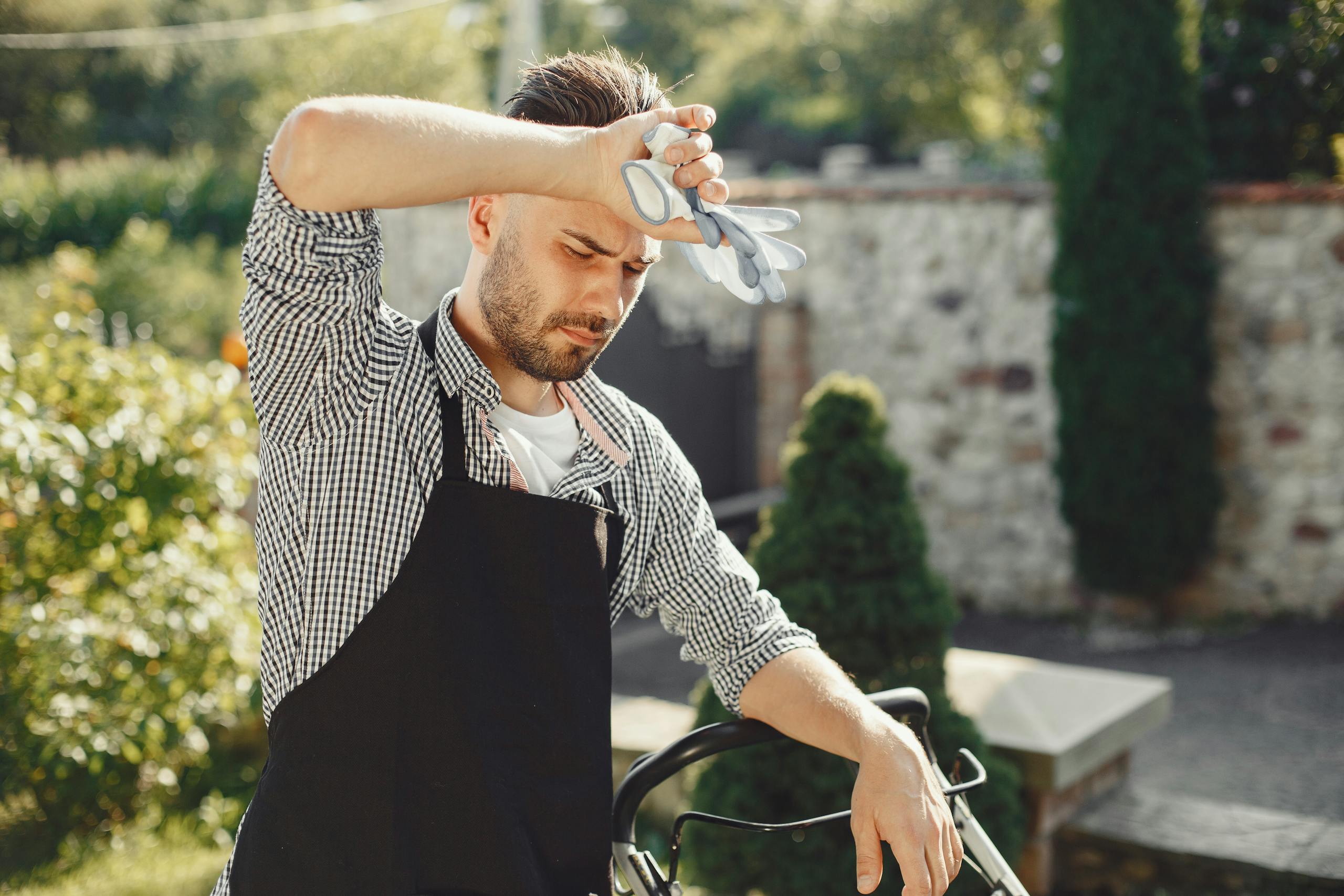 A tired gardener wipes sweat, resting from lawn care in a sunny backyard.