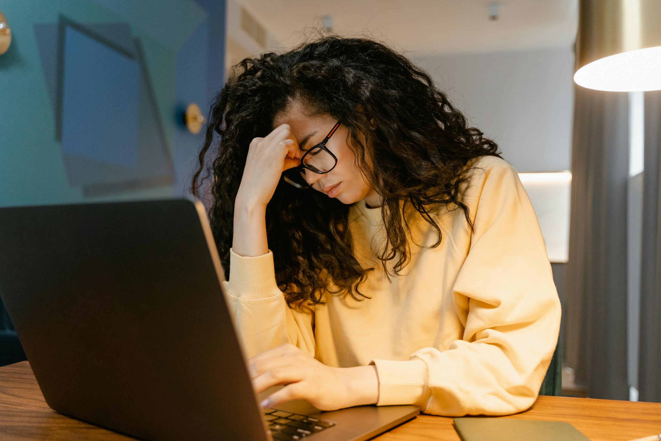 A tired woman with curly hair and glasses experiencing stress while working on a laptop.