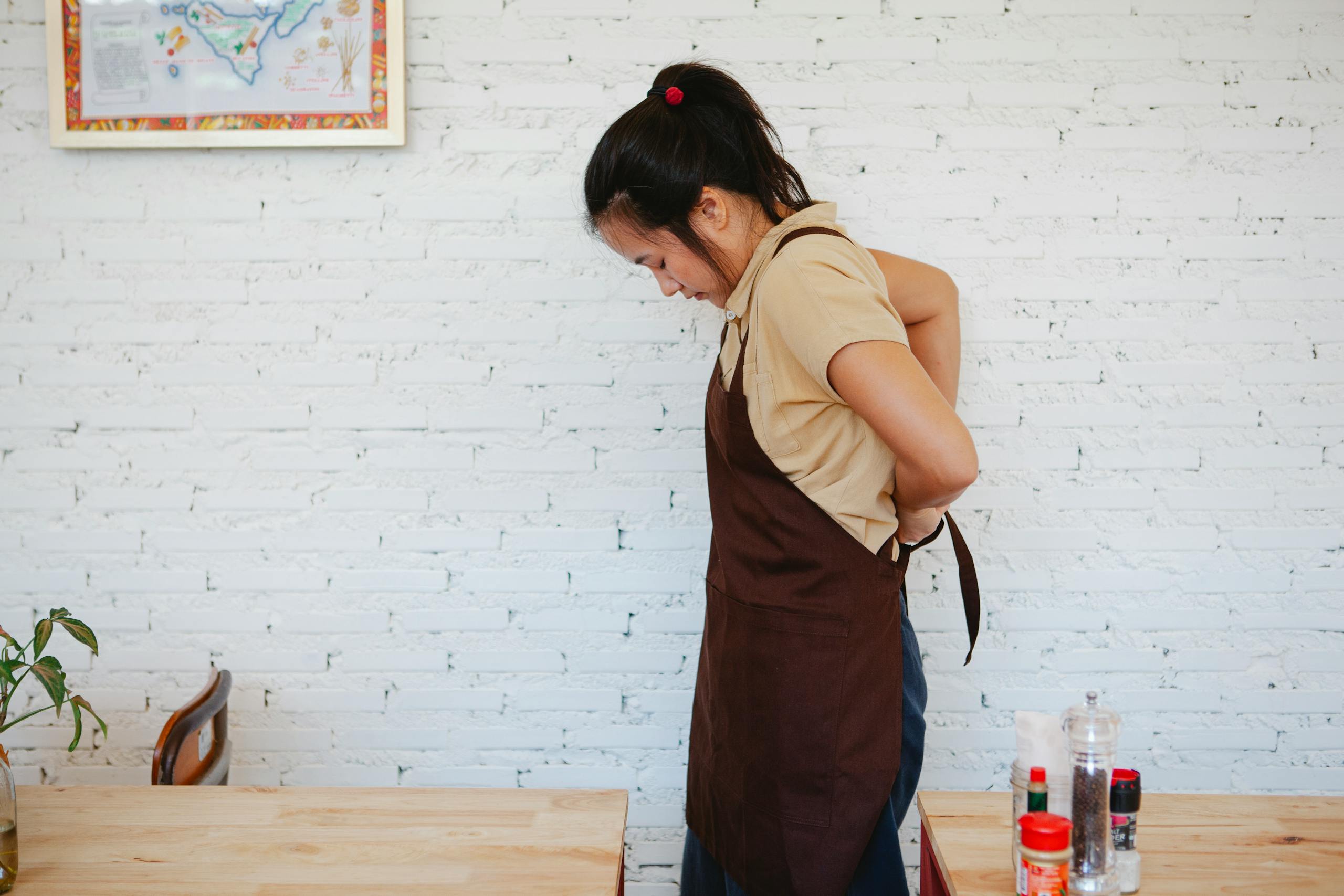 A waitress adjusts her apron inside a café with a white brick wall background.
