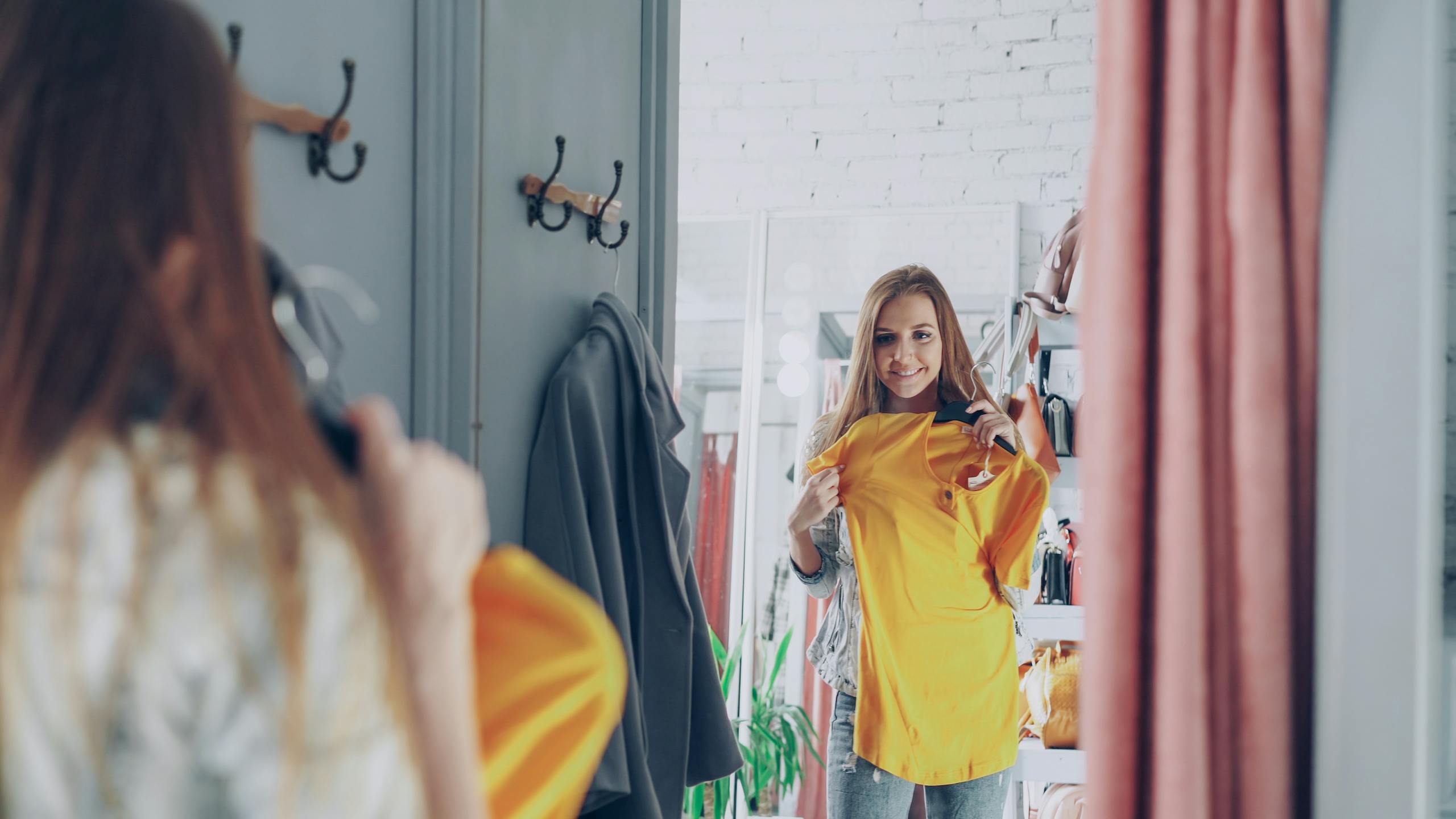 A woman admires herself in a boutique mirror holding a yellow top, shopping for fashion apparel.