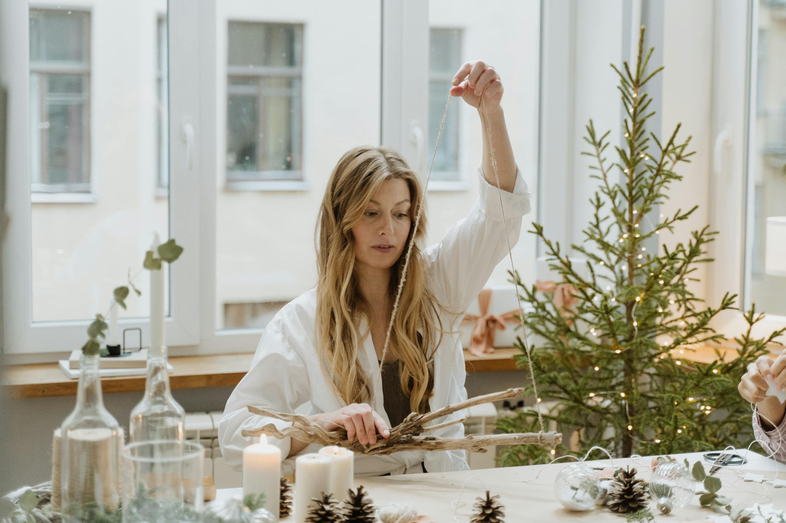 A woman decorates a room for Christmas with handmade ornaments near a small pine tree.