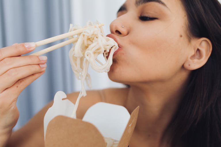 A woman enjoys Asian noodles using chopsticks in a takeout container.