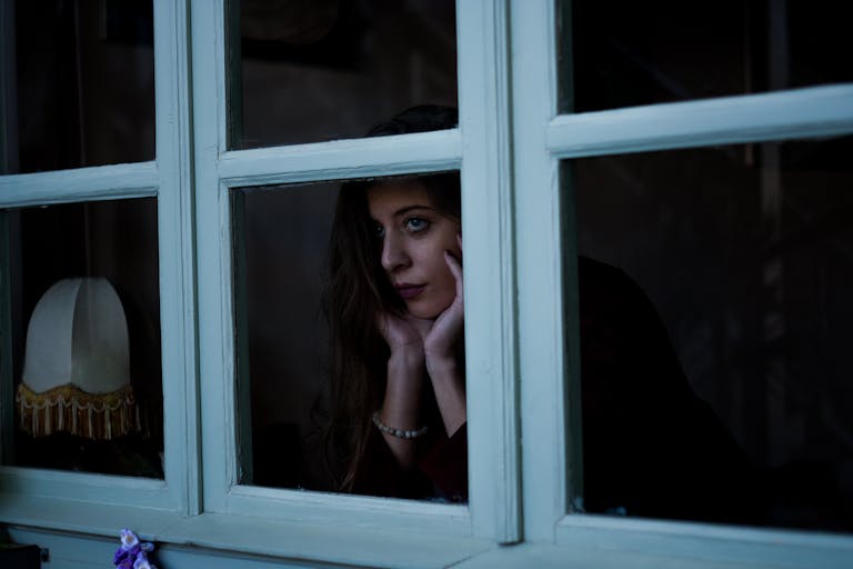 A woman gazes thoughtfully through a window at night, creating a moody and introspective scene.