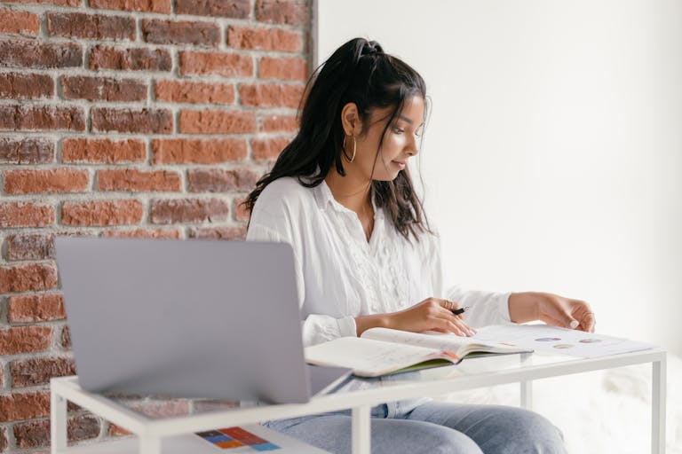 A woman in casual attire studies with a laptop and workbook at a desk indoors, beside a brick wall.