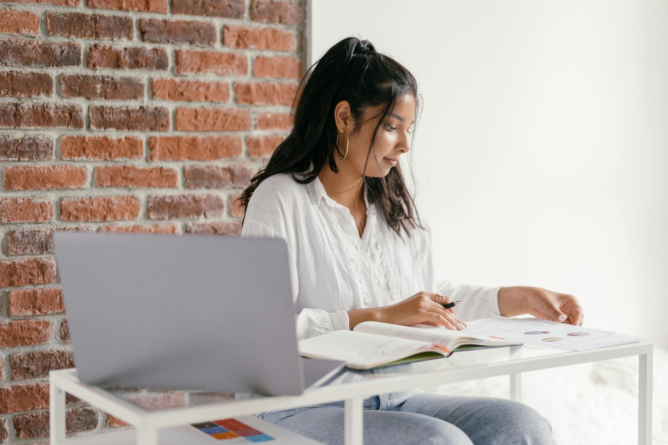 A woman in casual attire studies with a laptop and workbook at a desk indoors, beside a brick wall.