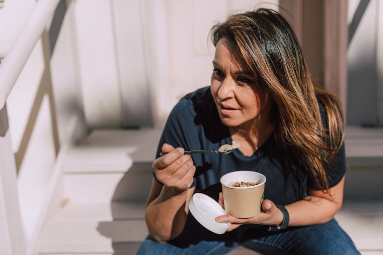 A woman in casual wear eating oats from a disposable bowl while sitting outdoors. Healthy lifestyle concept.