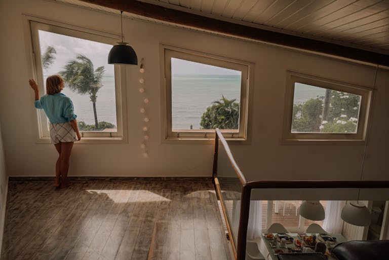 A woman looks out at the ocean view from a stylish modern home interior with large windows.