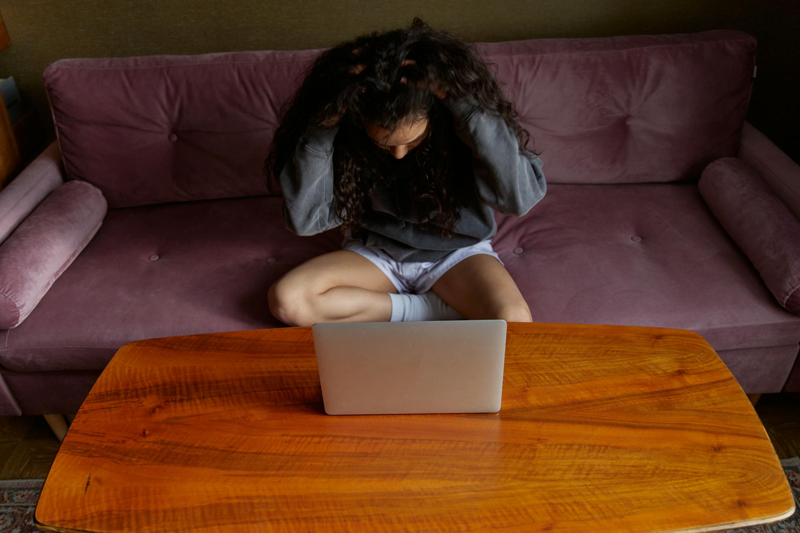 A woman on a sofa, stressed and holding her head over a laptop. Indoors setting.