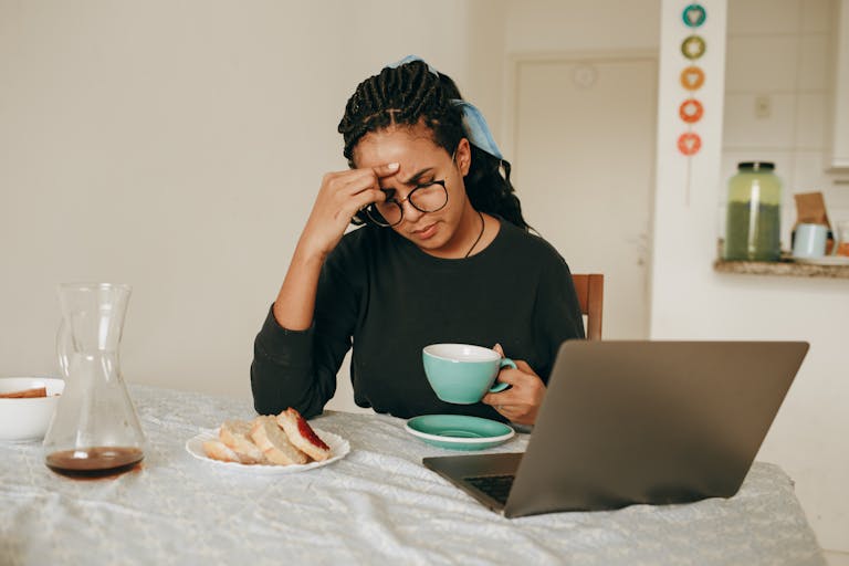 A woman sits at a dining table, holding a cup of coffee, appearing stressed while working on a laptop.