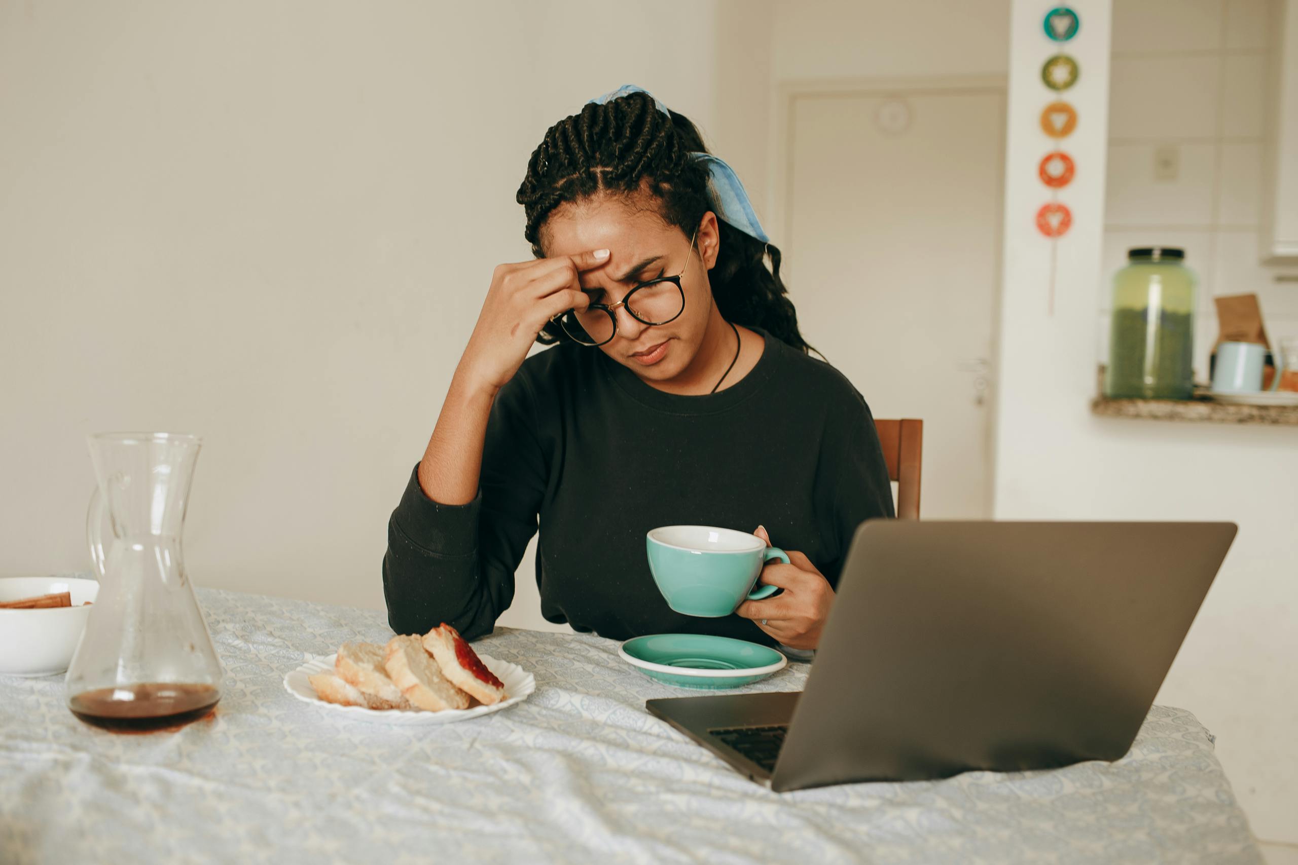A woman sits at a dining table, holding a cup of coffee, appearing stressed while working on a laptop.