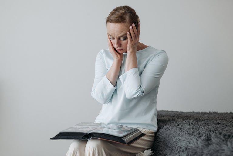 A woman sits thoughtfully with her hands on her face, looking at a photo album.