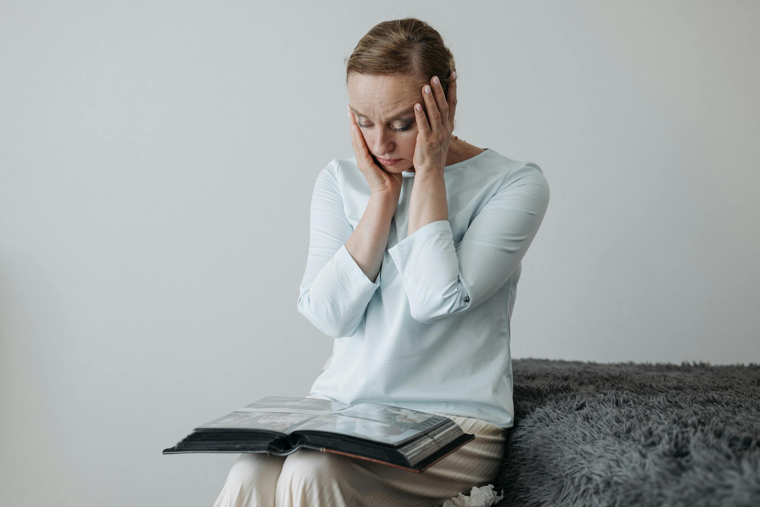 A woman sits thoughtfully with her hands on her face, looking at a photo album.