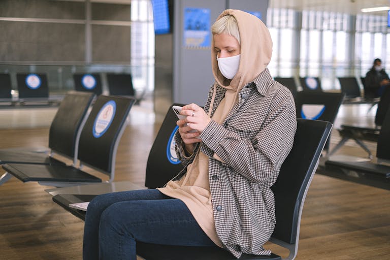 A woman wearing a face mask and a hood sits in an airport waiting area, using her smartphone.