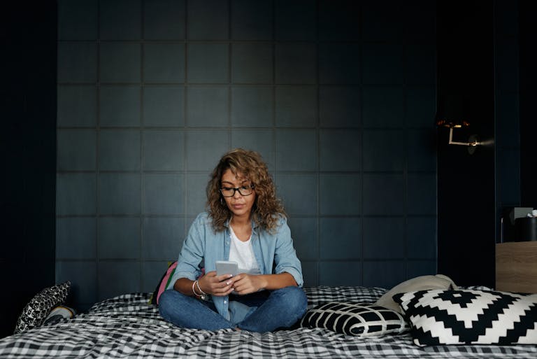A woman wearing glasses uses her smartphone, sitting on a bed in a cozy bedroom.