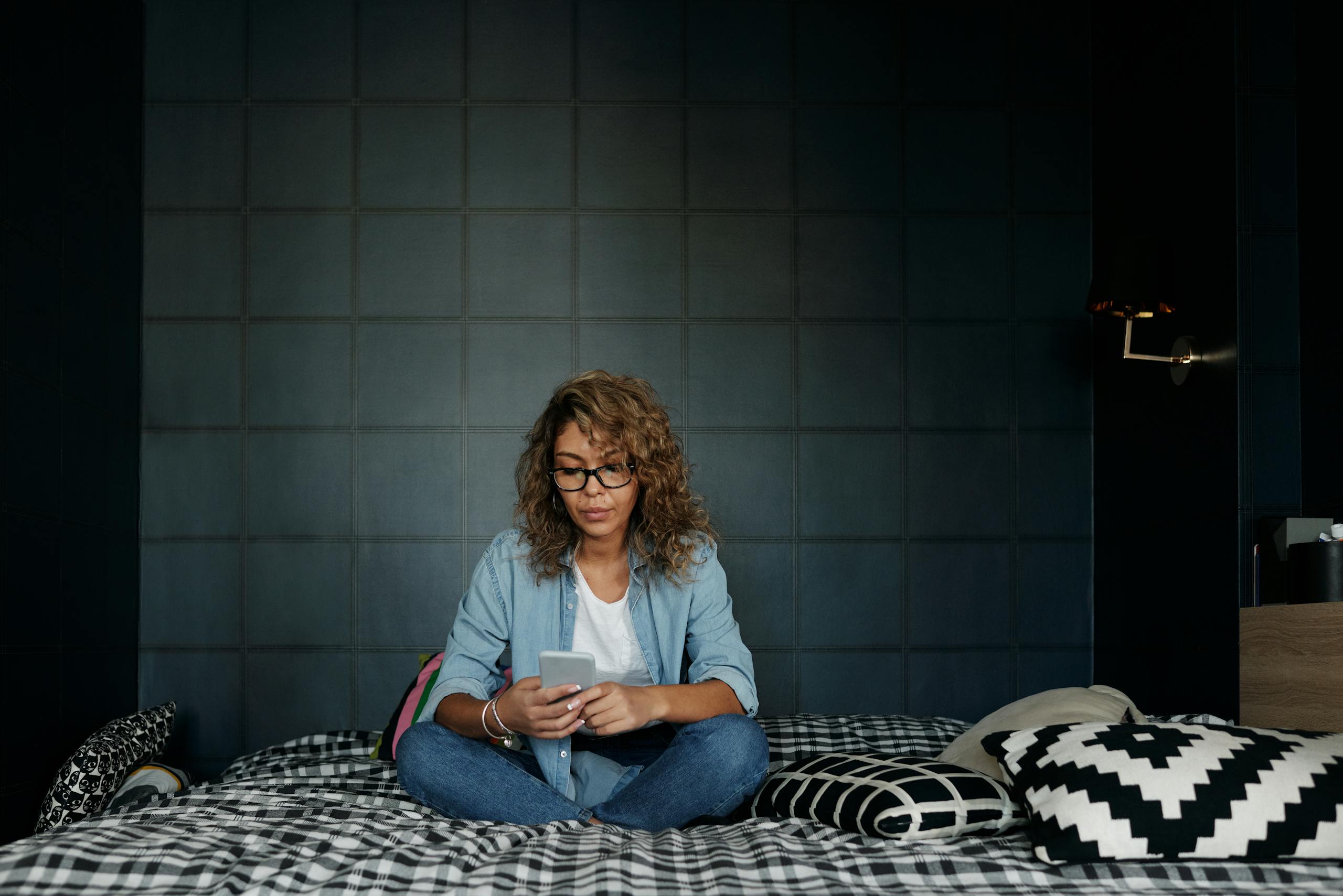 A woman wearing glasses uses her smartphone, sitting on a bed in a cozy bedroom.