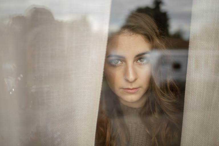 A woman with a nose piercing looks thoughtfully through a window, capturing a contemplative mood.
