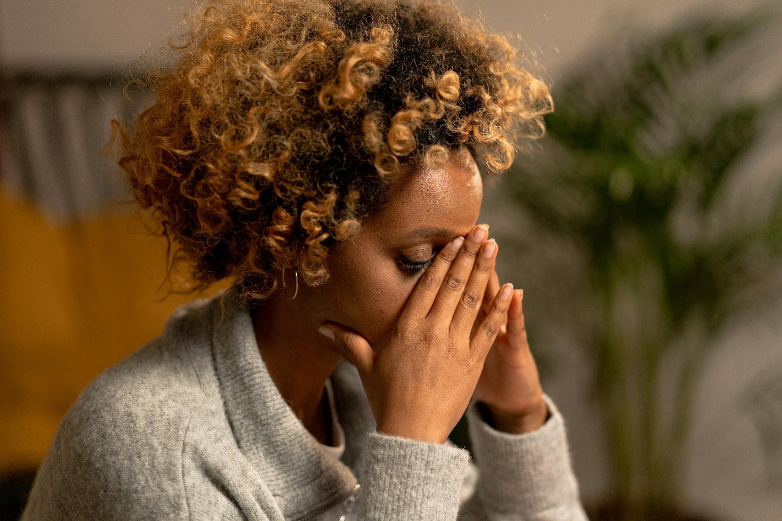 A woman with curly hair in a gray sweater, holding her face, depicting stress indoors.