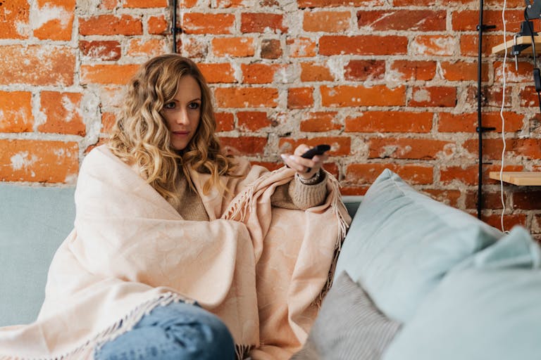 A woman wrapped in a blanket holding a remote control while sitting on a sofa indoors.