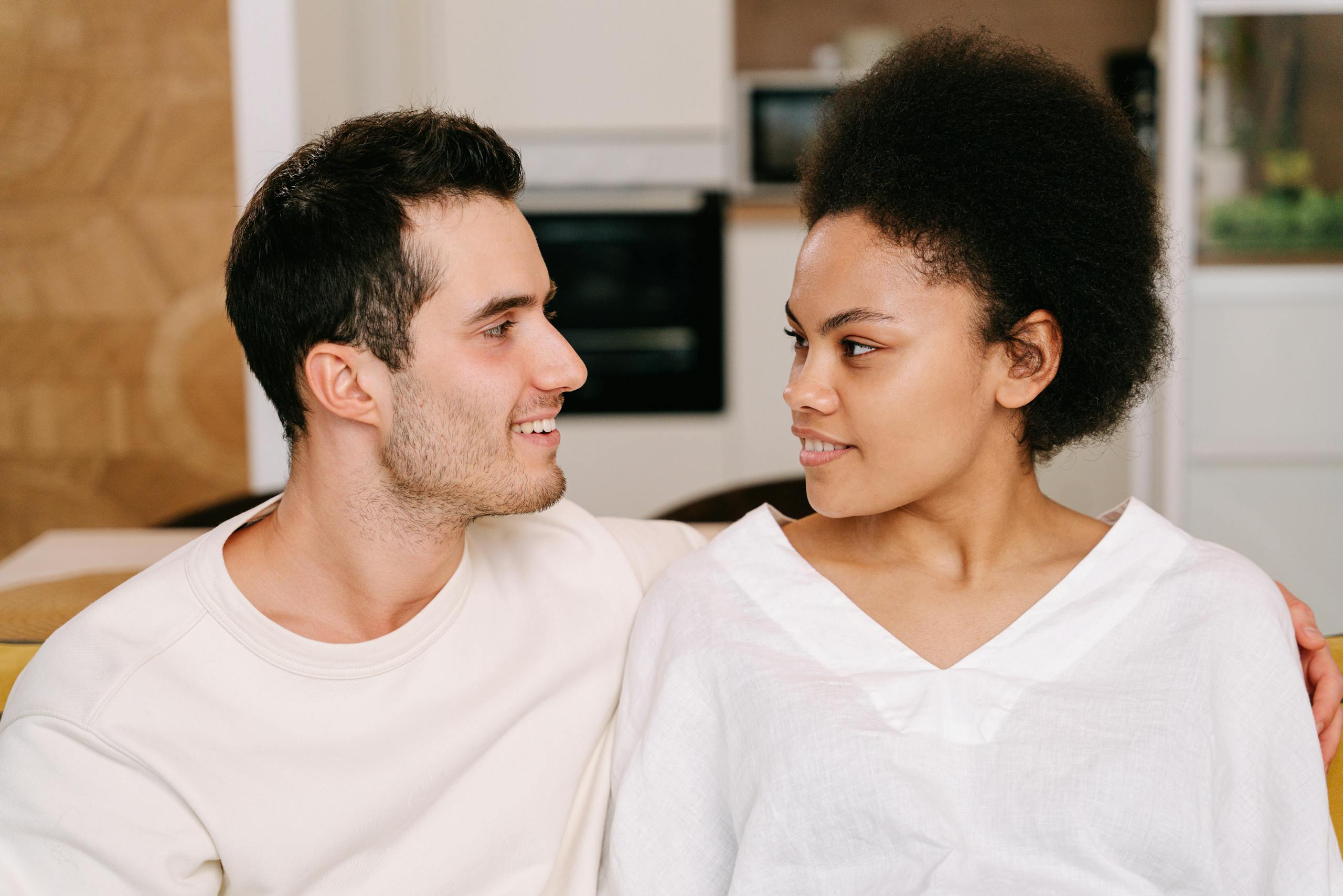 A young interracial couple sitting close together inside a cozy home setting, smiling warmly at each other.