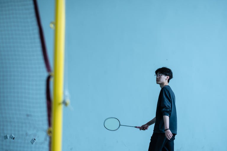 A young man holding a badminton racket standing indoors by the net.