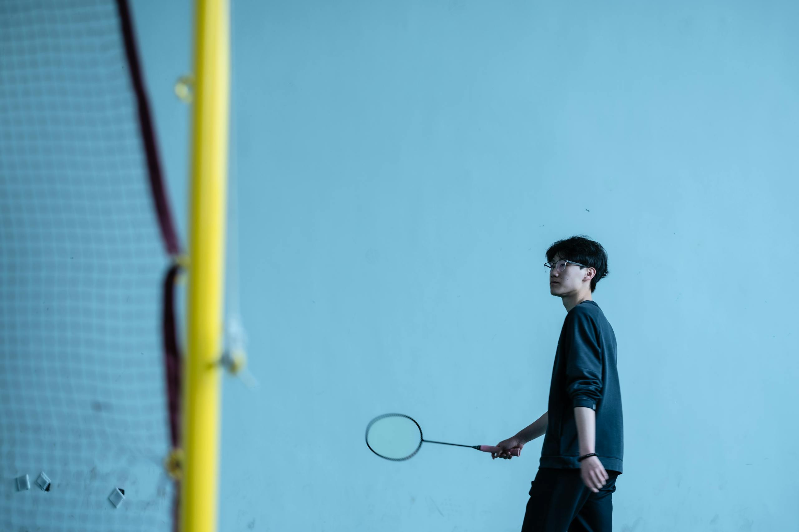 A young man holding a badminton racket standing indoors by the net.