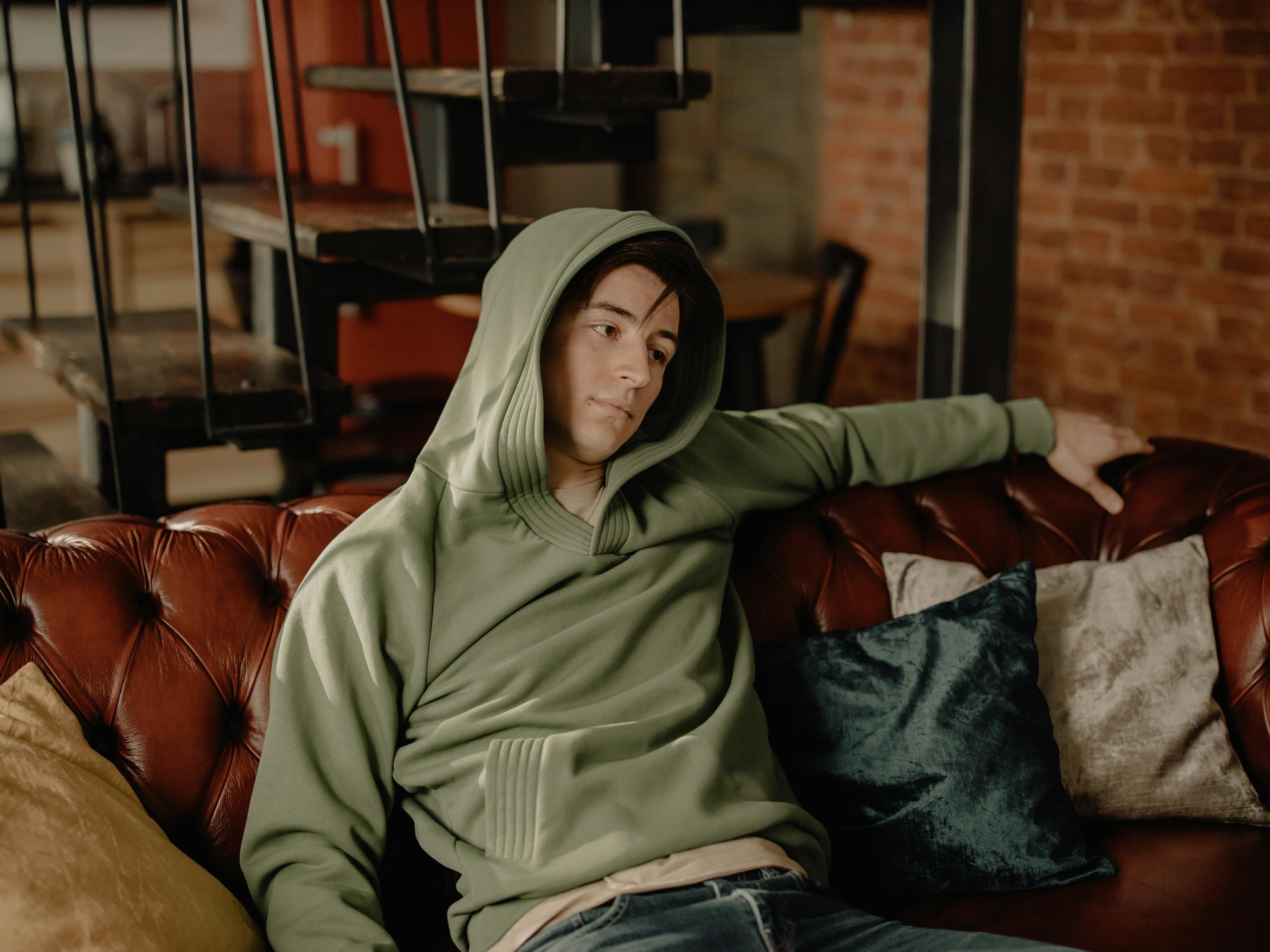 A young man wearing a green hoodie lounges on a sofa in a cozy home interior.
