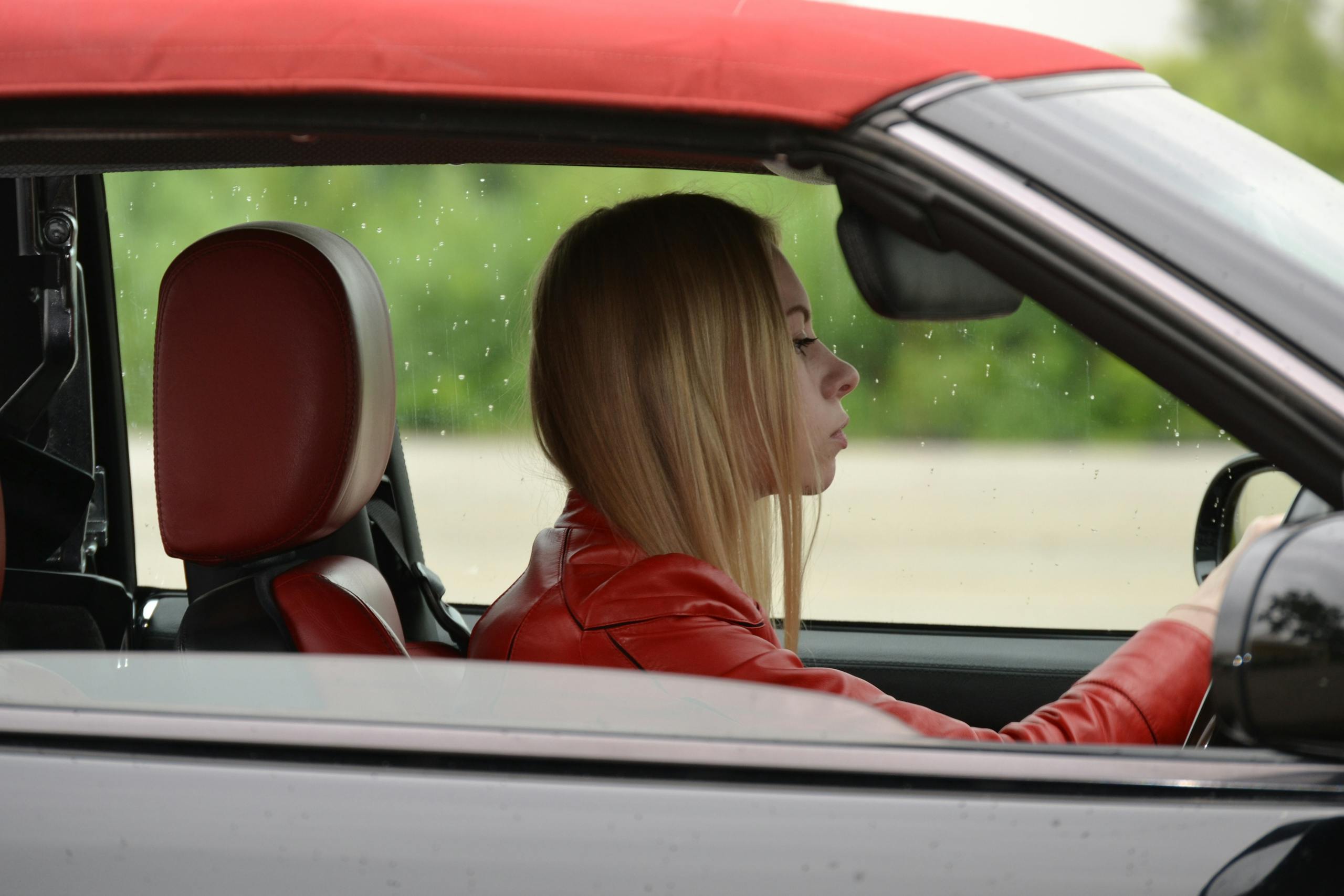 A young woman in a red convertible driving on a rainy day, wearing a red jacket.