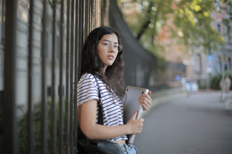 A young woman with glasses and striped shirt holding a tablet outdoors.