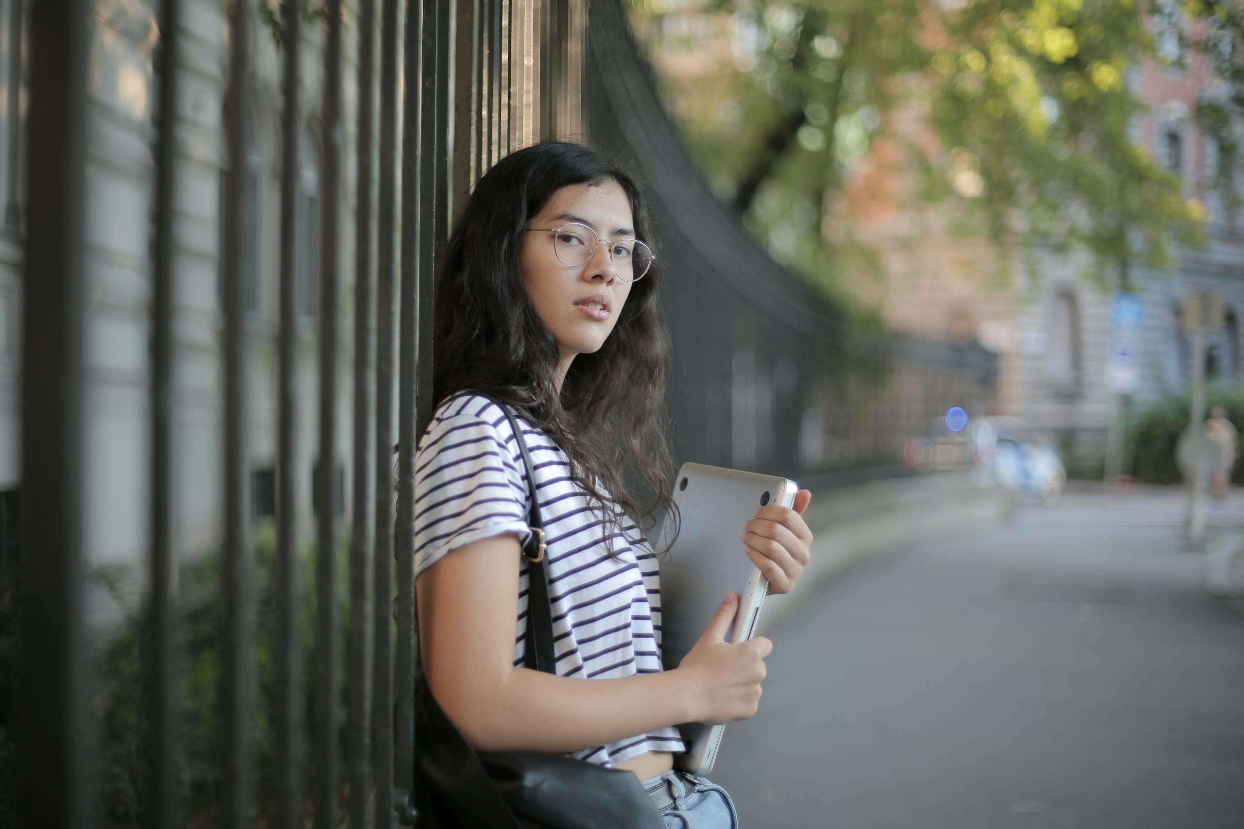 A young woman with glasses and striped shirt holding a tablet outdoors.
