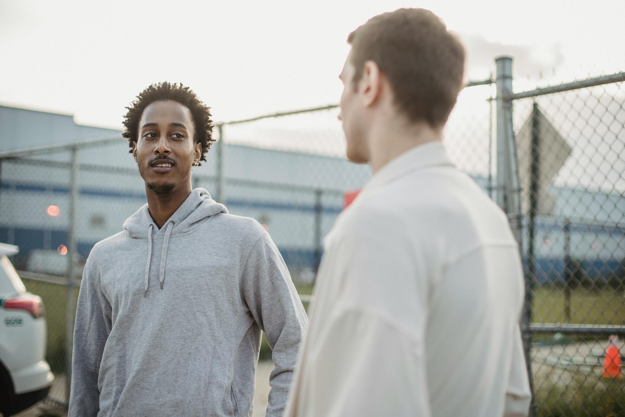 African American male standing with friend against metal grid near car on parking lot