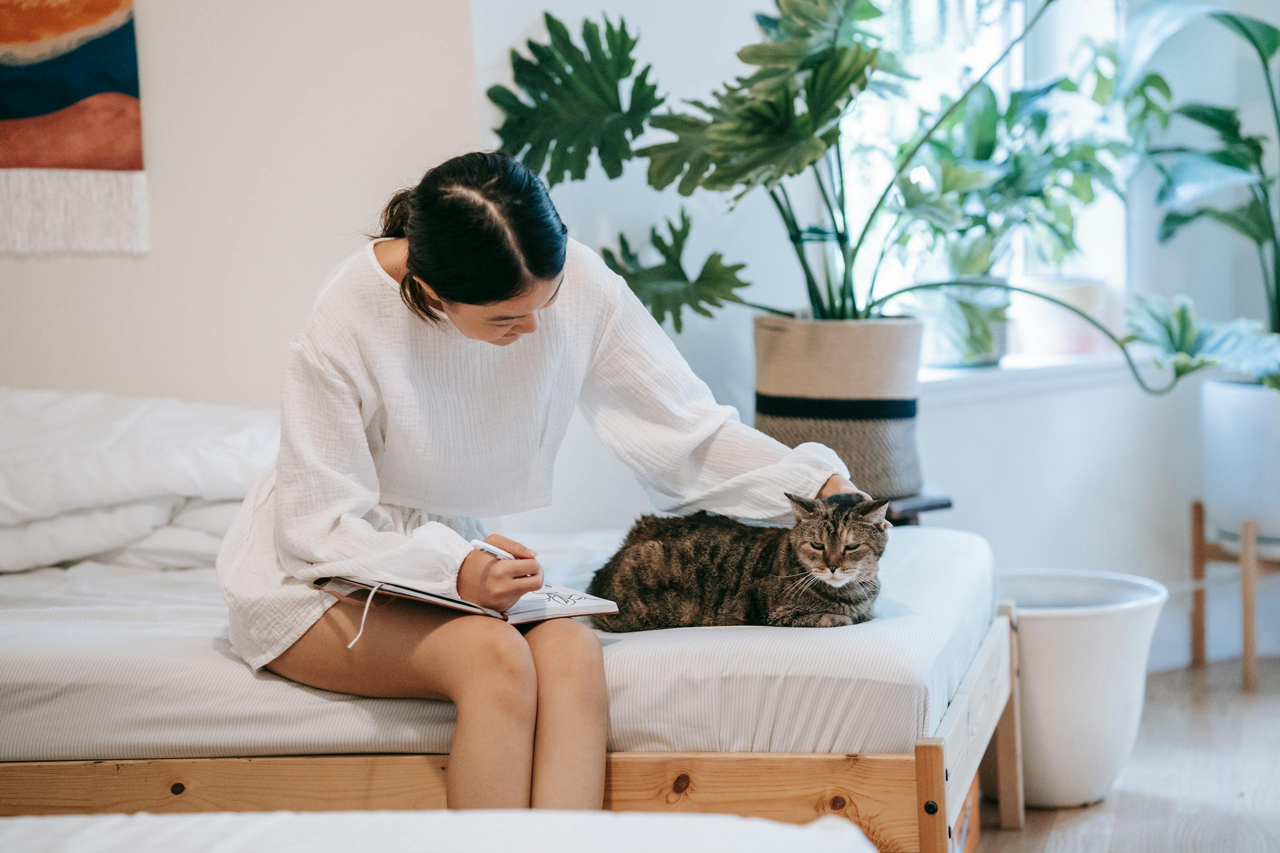 An Asian woman in a white outfit journaling while petting her cat on a bed surrounded by plants.