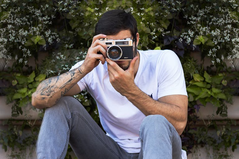 Anonymous tattooed male in stylish apparel taking photo on digital camera while sitting near blooming shrub
